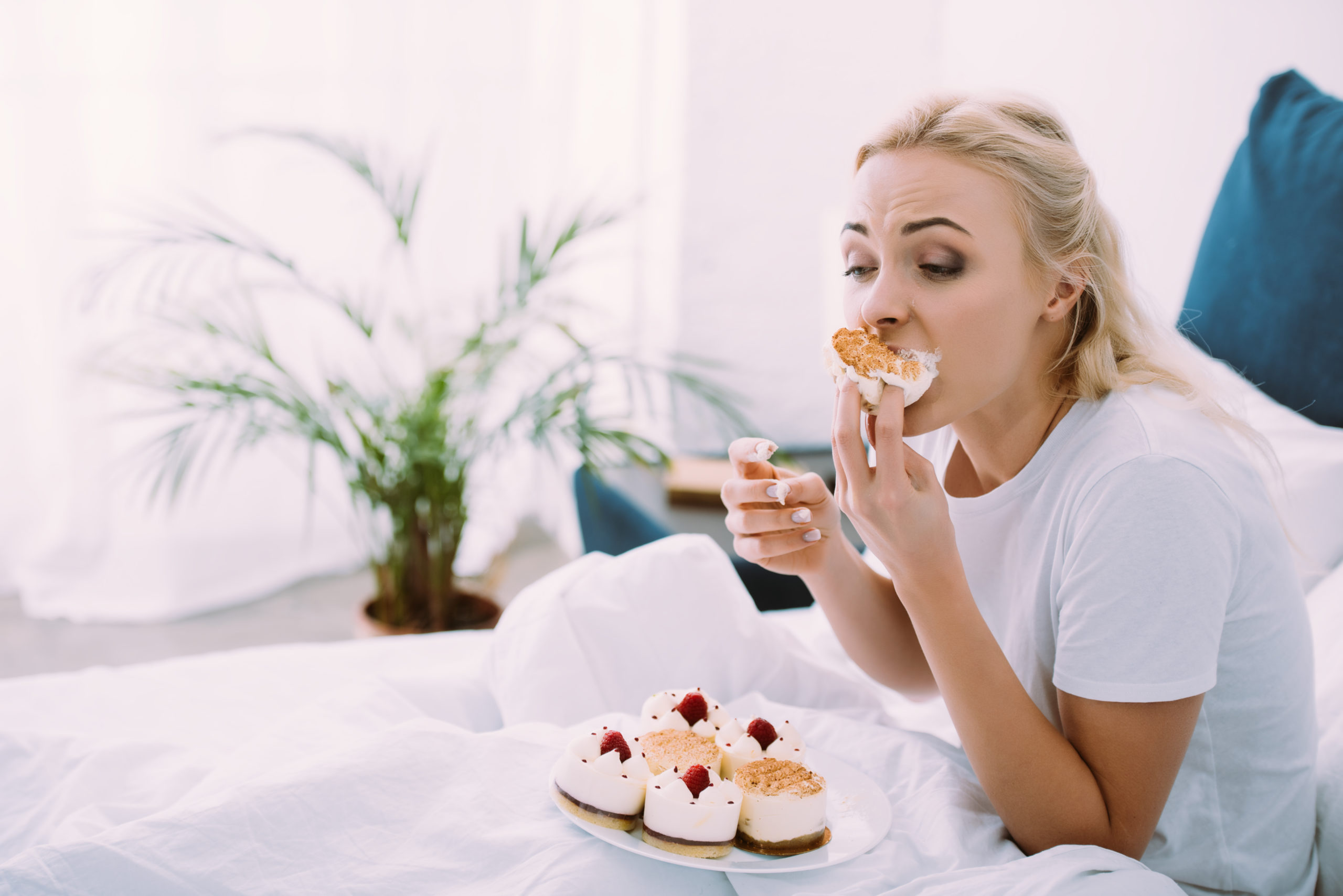 Blonde woman eating desserts in bed with a plate of pastries